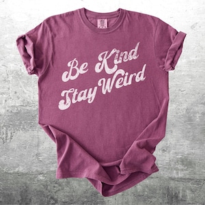May include: A maroon-colored t-shirt with the words "Be Kind Stay Weird" in a distressed white font. The shirt is slightly wrinkled and has rolled-up sleeves, suggesting a casual style. The background is a textured gray surface.