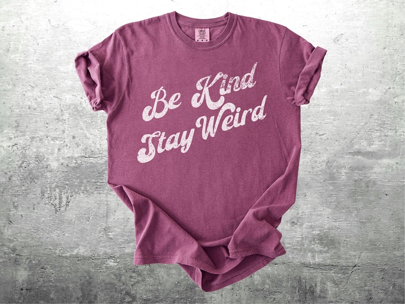 May include: A maroon-colored t-shirt with the words "Be Kind Stay Weird" in a distressed white font. The shirt is slightly wrinkled and has rolled-up sleeves, suggesting a casual style. The background is a textured gray surface.
