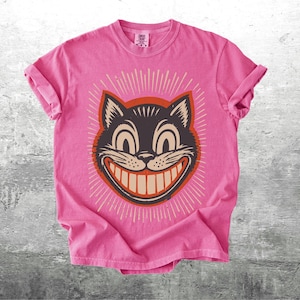 Op de afbeelding: Een felroze t-shirt met korte mouwen en een afbeelding van een zwart kattengezicht met een brede, tandige grijns. De kat is omlijst door een donkeroranje cirkel en uitstralende witte lijnen. Het shirt heeft een ronde hals en een casual pasvorm.