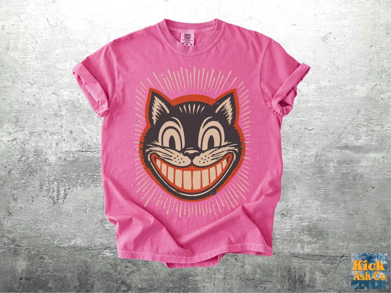 Op de afbeelding: Een felroze t-shirt met korte mouwen en een afbeelding van een zwart kattengezicht met een brede, tandige grijns. De kat is omlijst door een donkeroranje cirkel en uitstralende witte lijnen. Het shirt heeft een ronde hals en een casual pasvorm.