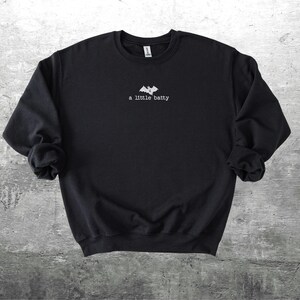 May include: Black crewneck sweatshirt with the phrase "a little batty" in white script below a bat graphic. The sweatshirt is made of a soft material and is suitable for casual wear.
