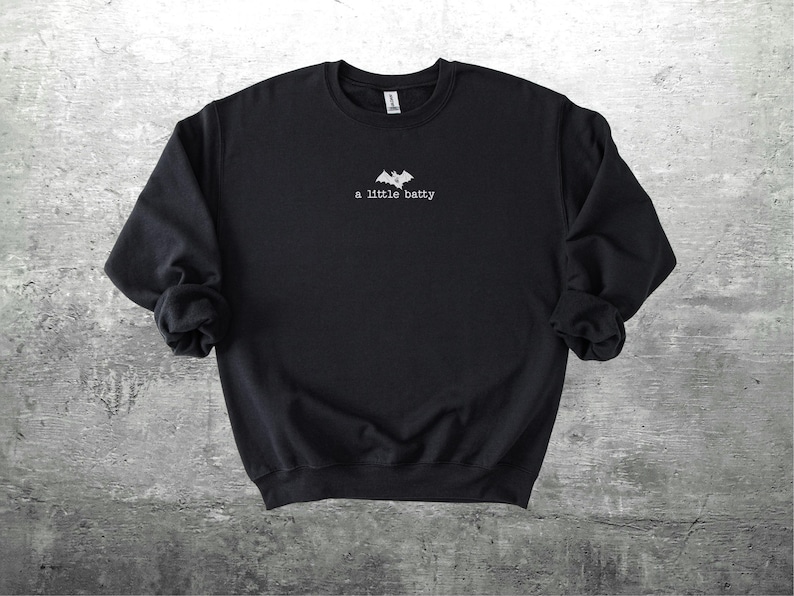 May include: Black crewneck sweatshirt with the phrase "a little batty" in white script below a bat graphic. The sweatshirt is made of a soft material and is suitable for casual wear.