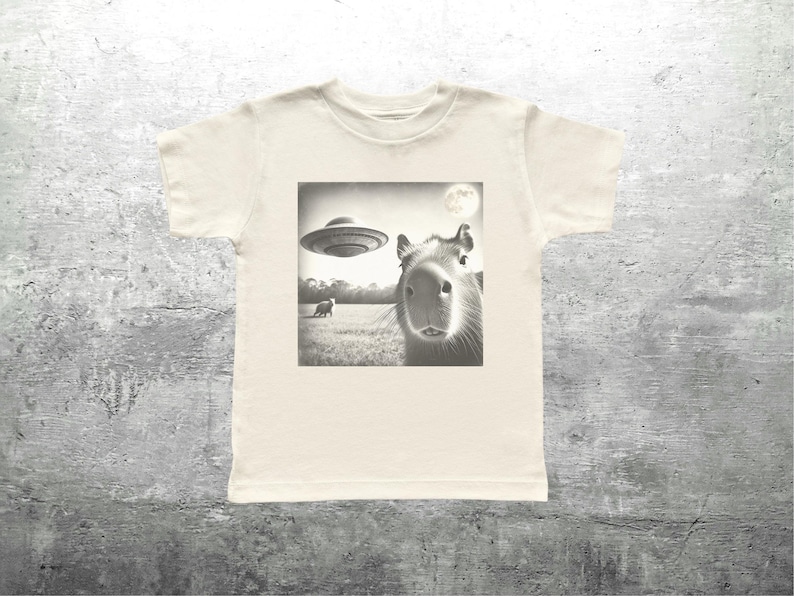 May include: A white t-shirt with a black and white image of a capybara looking up at a flying saucer. The image also shows a small animal in the distance.