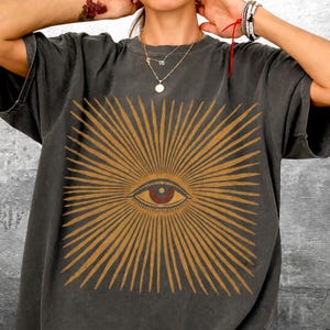 May include: Dark gray t-shirt featuring a gold sunburst design with a central eye graphic. The eye has a red iris and is detailed with fine lines. The shirt has a vintage, worn look.