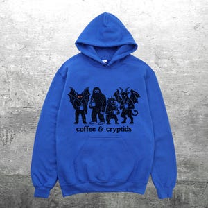 May include: A royal blue hooded sweatshirt with a front pocket and a black graphic. The graphic features four cryptid figures holding coffee mugs, with the words "coffee & cryptids" printed below.