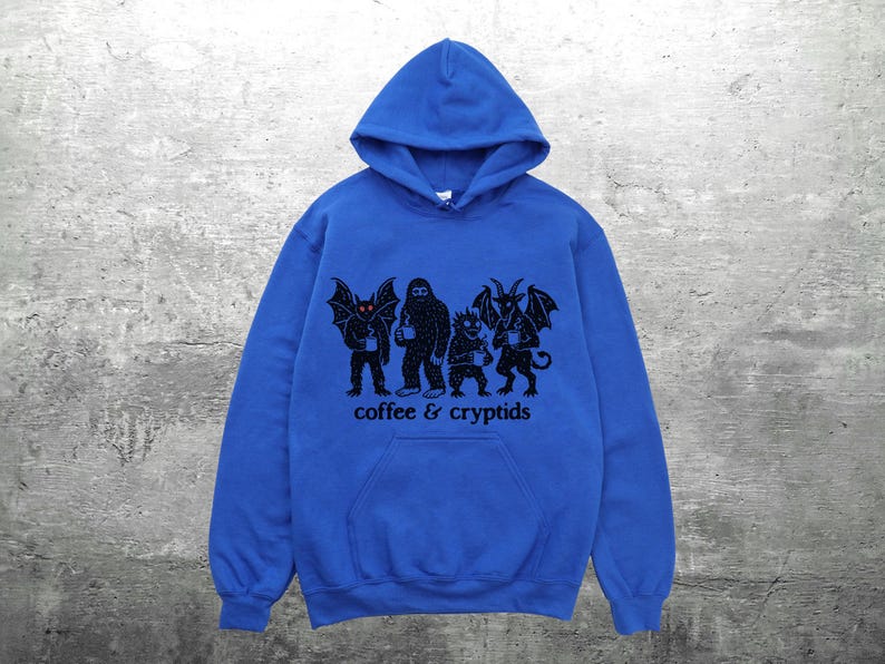 May include: A royal blue hooded sweatshirt with a front pocket and a black graphic. The graphic features four cryptid figures holding coffee mugs, with the words "coffee & cryptids" printed below.