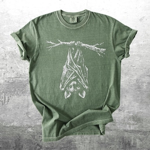 Op de afbeelding: Olijfgroen t-shirt met een witte illustratie van een vleermuis die aan een tak hangt. Het t-shirt heeft opgerolde mouwen en een ronde hals. Het vleermuisontwerp staat in het midden van de voorkant.