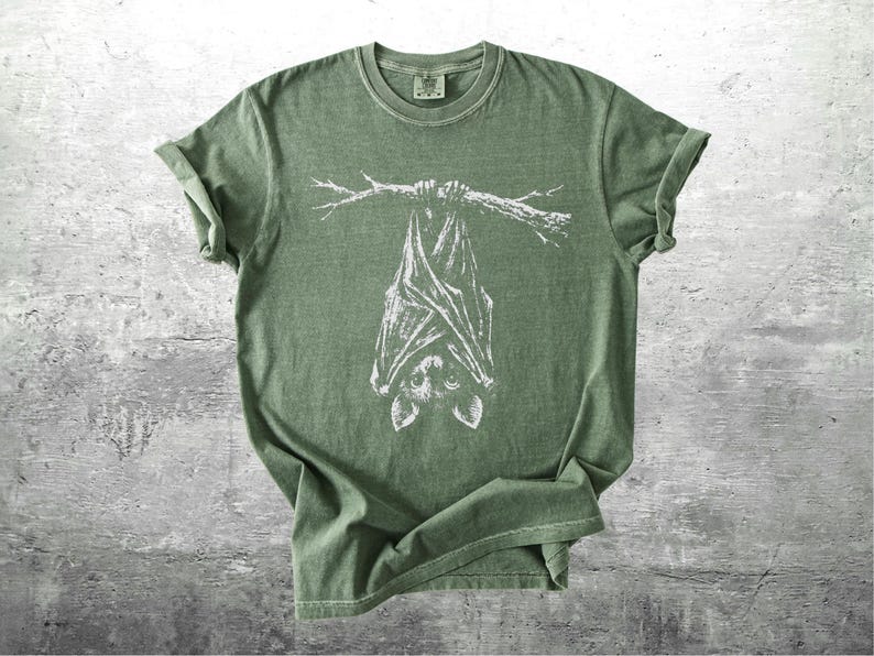 Op de afbeelding: Olijfgroen t-shirt met een witte illustratie van een vleermuis die aan een tak hangt. Het t-shirt heeft opgerolde mouwen en een ronde hals. Het vleermuisontwerp staat in het midden van de voorkant.