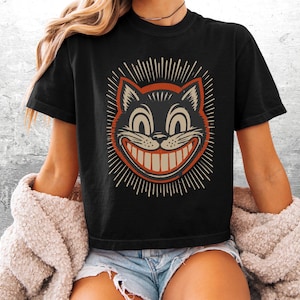 May include: A black short-sleeve t-shirt featuring a graphic of a grinning cat face with large eyes and a wide smile. The cat illustration is outlined in orange and surrounded by radiating white lines. The shirt has a crew neckline and a boxy fit.