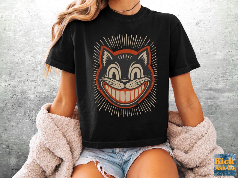 May include: A black short-sleeve t-shirt featuring a graphic of a grinning cat face with large eyes and a wide smile. The cat illustration is outlined in orange and surrounded by radiating white lines. The shirt has a crew neckline and a boxy fit.