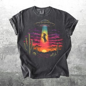 May include: A charcoal gray short-sleeve t-shirt featuring a graphic print of a dachshund being abducted by a UFO in a desert landscape at sunset. The design includes cacti, a bright sun, and a colorful beam of light against a dark sky.