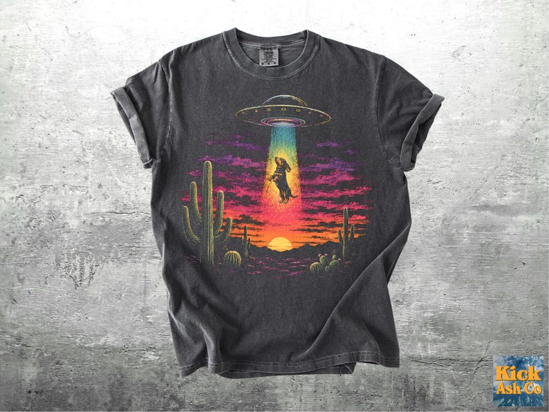 May include: A charcoal gray short-sleeve t-shirt featuring a graphic print of a dachshund being abducted by a UFO in a desert landscape at sunset. The design includes cacti, a bright sun, and a colorful beam of light against a dark sky.