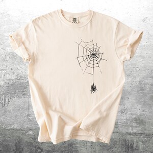 May include: Cream-colored t-shirt featuring a black spider web design with a spider hanging below. The t-shirt has short sleeves and a crew neck. The fabric is soft and appears to be cotton.