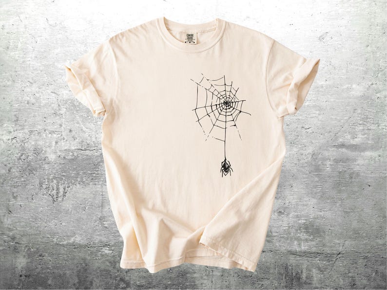 May include: Cream-colored t-shirt featuring a black spider web design with a spider hanging below. The t-shirt has short sleeves and a crew neck. The fabric is soft and appears to be cotton.