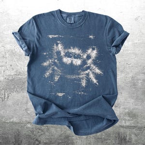 Op de afbeelding: Een blauw t-shirt met een witte, verweerde afbeelding van een springspin. Het shirt met korte mouwen is op een grijze ondergrond gevouwen. Het spinnenontwerp heeft gedetailleerde ogen en poten.
