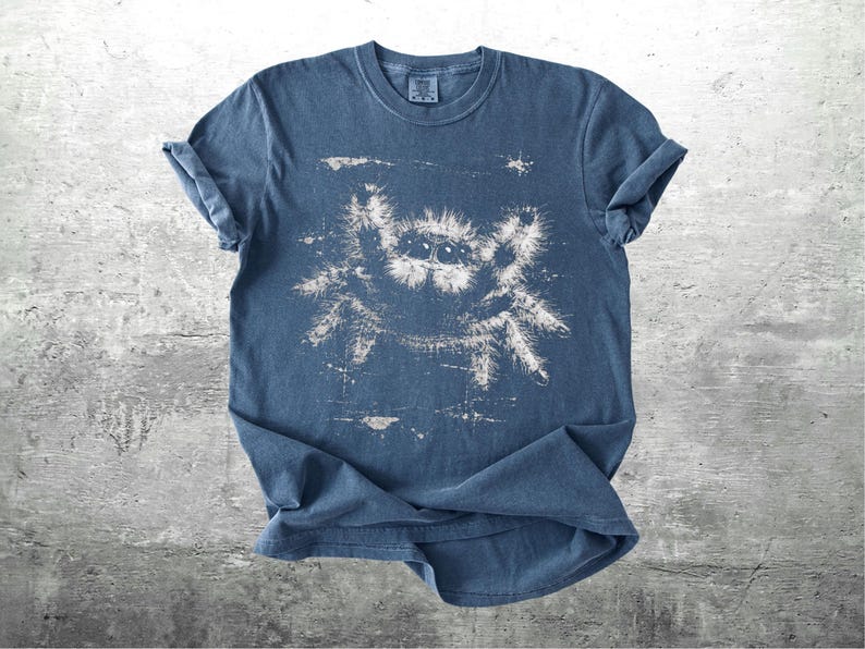Op de afbeelding: Een blauw t-shirt met een witte, verweerde afbeelding van een springspin. Het shirt met korte mouwen is op een grijze ondergrond gevouwen. Het spinnenontwerp heeft gedetailleerde ogen en poten.