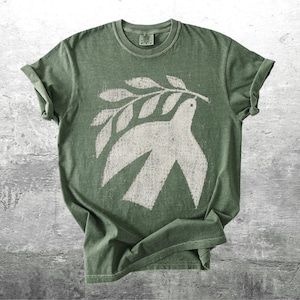 May include: A sage green short-sleeve t-shirt featuring a distressed white graphic print of a dove carrying an olive branch. The shirt has a crew neckline and a vintage-style wash, with the text Kick Ash Co. visible in the bottom right corner.