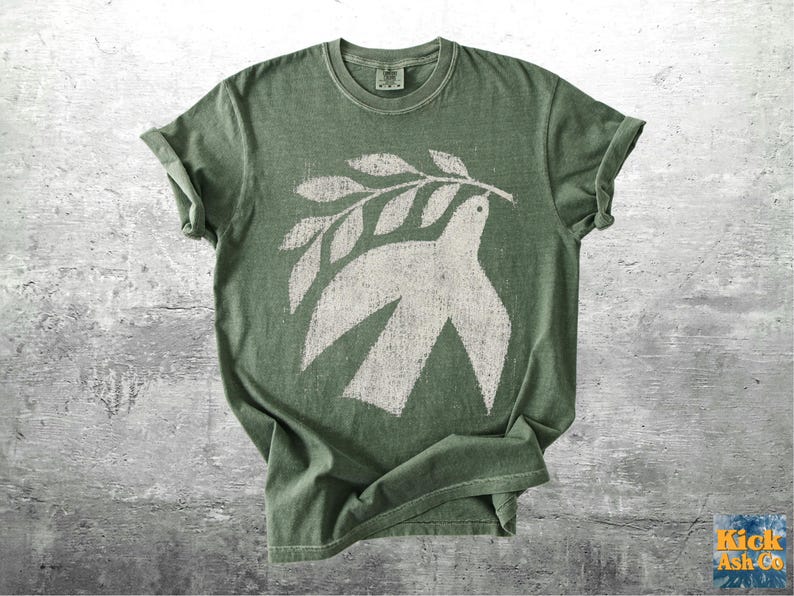 May include: A sage green short-sleeve t-shirt featuring a distressed white graphic print of a dove carrying an olive branch. The shirt has a crew neckline and a vintage-style wash, with the text Kick Ash Co. visible in the bottom right corner.