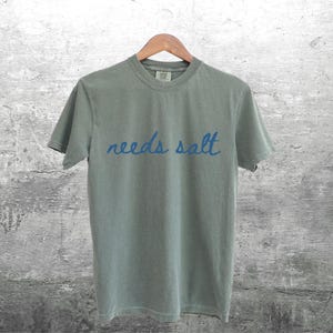 May include: A light gray t-shirt with the text "needs salt" written in blue cursive.