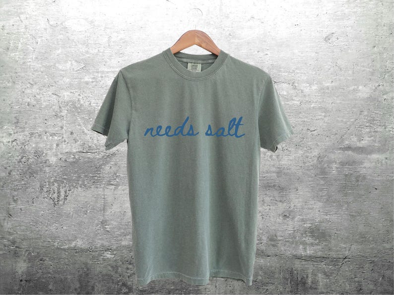 May include: A light gray t-shirt with the text "needs salt" written in blue cursive.