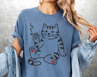 May include: Blue t-shirt featuring a cartoon cat with a cigarette. The cat is sitting with its legs crossed, and has black stripes and red heart details on its paws. The cat is smoking a cigarette.