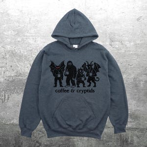 May include: A dark gray hooded sweatshirt with a front pocket and a drawstring hood. The sweatshirt features a black graphic of four cryptids holding coffee cups, with the words "coffee & cryptids" printed below.