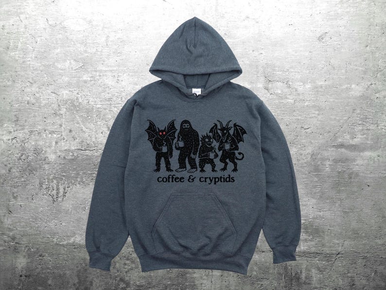 May include: A dark gray hooded sweatshirt with a front pocket and a drawstring hood. The sweatshirt features a black graphic of four cryptids holding coffee cups, with the words "coffee & cryptids" printed below.