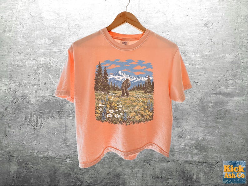 Puede incluir: Camiseta color melocot&oacute;n con un gr&aacute;fico de un Bigfoot en un campo con &aacute;rboles, monta&ntilde;as y cielo azul. La camiseta est&aacute; colgada en una percha de madera contra una pared de hormig&oacute;n gris.