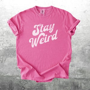 K&ouml;nnte beinhalten: Ein pinkfarbenes T-Shirt mit dem wei&szlig;en Schriftzug "Stay Weird" in kursiver Schrift. Das Shirt ist gefaltet, die &Auml;rmel sind hochgekrempelt. Der Hintergrund ist eine graue, strukturierte Oberfl&auml;che.