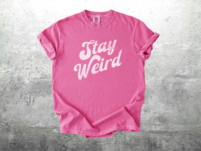K&ouml;nnte beinhalten: Ein pinkfarbenes T-Shirt mit dem wei&szlig;en Schriftzug "Stay Weird" in kursiver Schrift. Das Shirt ist gefaltet, die &Auml;rmel sind hochgekrempelt. Der Hintergrund ist eine graue, strukturierte Oberfl&auml;che.