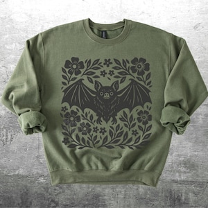 May include: Olive green sweatshirt with a black bat and floral design. The bat is centered within a square frame of flowers and leaves. The sweatshirt has long sleeves and a crew neck.