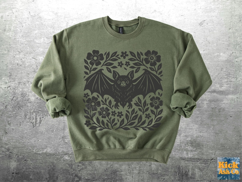 May include: Olive green sweatshirt with a black bat and floral design. The bat is centered within a square frame of flowers and leaves. The sweatshirt has long sleeves and a crew neck.