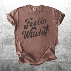 K&ouml;nnte beinhalten: Ein braunes T-Shirt mit dem Text "Feelin Witchy" in schwarzer, verwaschener Schrift und einer Hexenhut-Grafik. Das Shirt ist gefaltet, mit hochgekrempelten &Auml;rmeln, auf einer grauen Oberfl&auml;che.