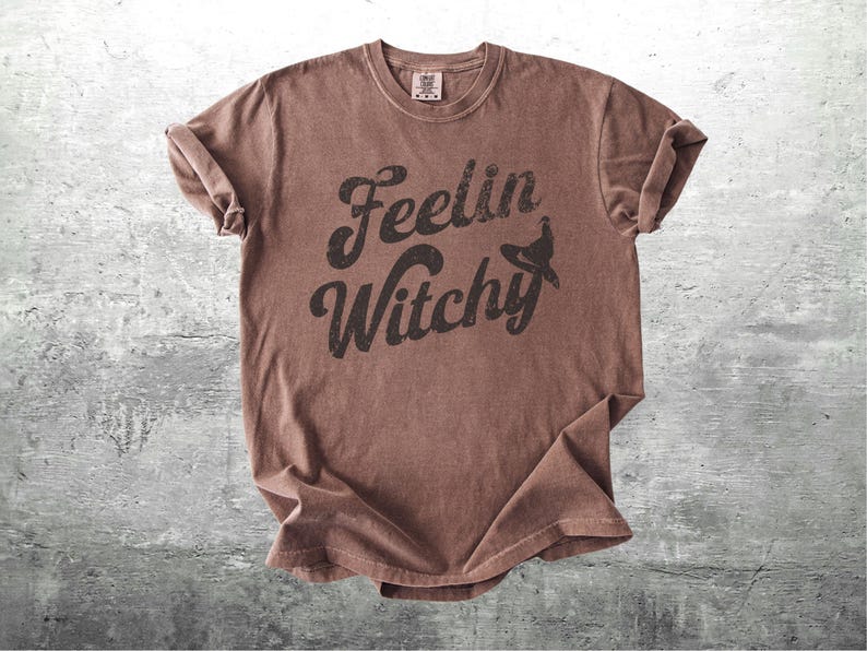K&ouml;nnte beinhalten: Ein braunes T-Shirt mit dem Text "Feelin Witchy" in schwarzer, verwaschener Schrift und einer Hexenhut-Grafik. Das Shirt ist gefaltet, mit hochgekrempelten &Auml;rmeln, auf einer grauen Oberfl&auml;che.