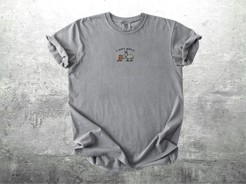 May include: A grey t-shirt with rolled sleeves. The shirt has a small graphic of a donkey and a carrot with the text "I don't give a". The t-shirt is made of a soft material and is suitable for casual wear.