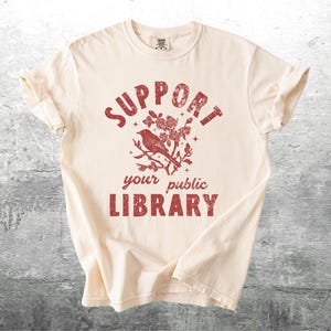 May include: Cream-colored t-shirt with a distressed red graphic. The graphic features the words "SUPPORT your public LIBRARY" surrounding a bird perched on a flowering branch. The shirt has short sleeves and a crew neck.
