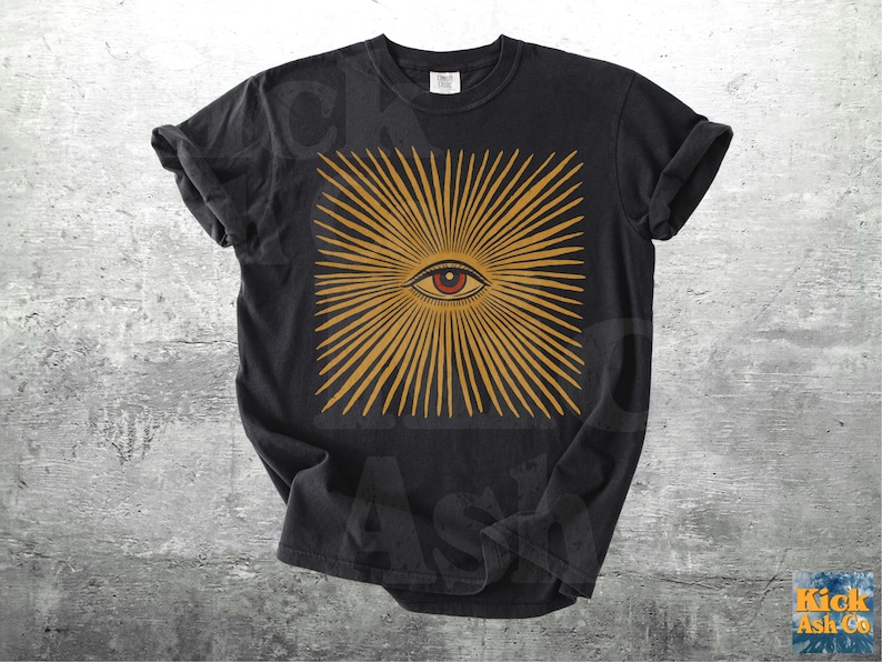 May include: Black t-shirt featuring a square design with a central eye and radiating golden lines. The eye is red and the design is centered on the shirt. The shirt is a classic crew neck style.