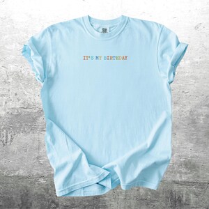 Puede incluir: Una camiseta azul claro con el texto "It's My Birthday" en colores arco&iacute;ris.