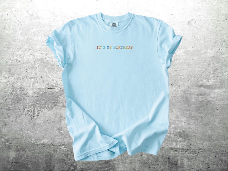 Puede incluir: Una camiseta azul claro con el texto "It's My Birthday" en colores arco&iacute;ris.