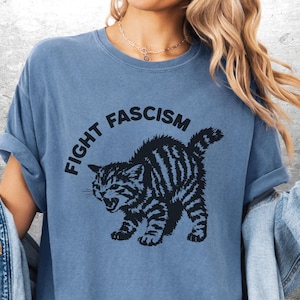 K&ouml;nnte beinhalten: Blaues T-Shirt mit dem Text "FIGHT FASCISM" &uuml;ber einer schwarz-wei&szlig; gestreiften Katzenillustration. Die Katze ist in einer aggressiven Haltung dargestellt. Die &Auml;rmel des T-Shirts sind hochgekrempelt.
