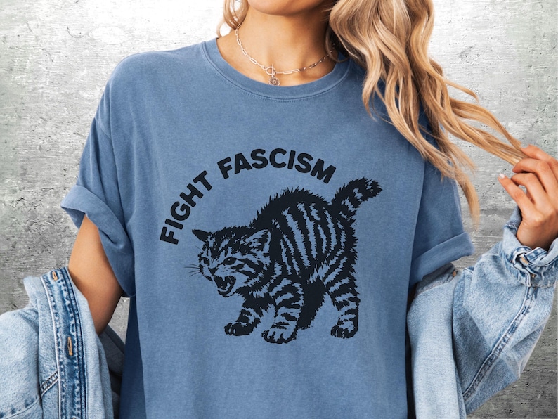 K&ouml;nnte beinhalten: Blaues T-Shirt mit dem Text "FIGHT FASCISM" &uuml;ber einer schwarz-wei&szlig; gestreiften Katzenillustration. Die Katze ist in einer aggressiven Haltung dargestellt. Die &Auml;rmel des T-Shirts sind hochgekrempelt.