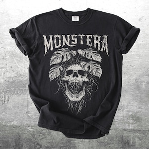 May include: Black t-shirt with a white graphic design. The design features the word "MONSTERA" in a gothic font above a skull with leaves. The shirt is made of a soft material and has rolled-up sleeves.