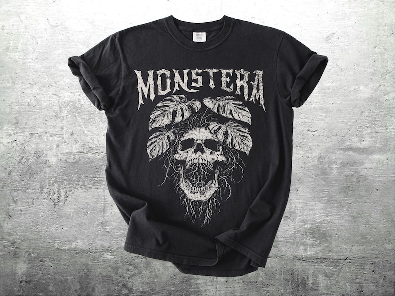 May include: Black t-shirt with a white graphic design. The design features the word "MONSTERA" in a gothic font above a skull with leaves. The shirt is made of a soft material and has rolled-up sleeves.