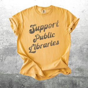 May include: Mustard yellow t-shirt with rolled-up sleeves. The shirt features the text "Support Public Libraries" in a distressed, dark gray font. The shirt is laid flat on a gray, textured surface.