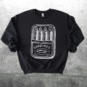 May include: Black sweatshirt with a white graphic of a sardine can. The can contains four sardines and the text "Sardines Wild Caught in Olive Oil". The design has a vintage, hand-drawn aesthetic.