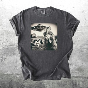 May include: Dark gray t-shirt featuring a black and white image of a chicken looking at a UFO hovering over a farm. The image includes other chickens and a barn. The shirt has short sleeves and a crew neck.