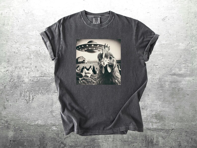 May include: Dark gray t-shirt featuring a black and white image of a chicken looking at a UFO hovering over a farm. The image includes other chickens and a barn. The shirt has short sleeves and a crew neck.