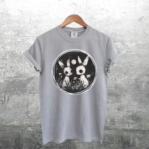 Op de afbeelding: Een grijs T-shirt met een zwart-witte afbeelding van twee konijnen met een volle maan op de achtergrond.