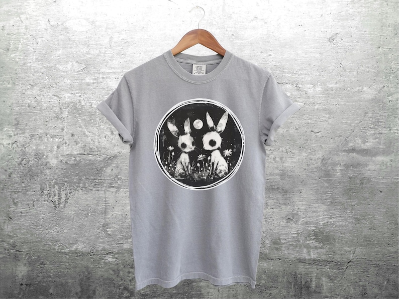 Op de afbeelding: Een grijs T-shirt met een zwart-witte afbeelding van twee konijnen met een volle maan op de achtergrond.