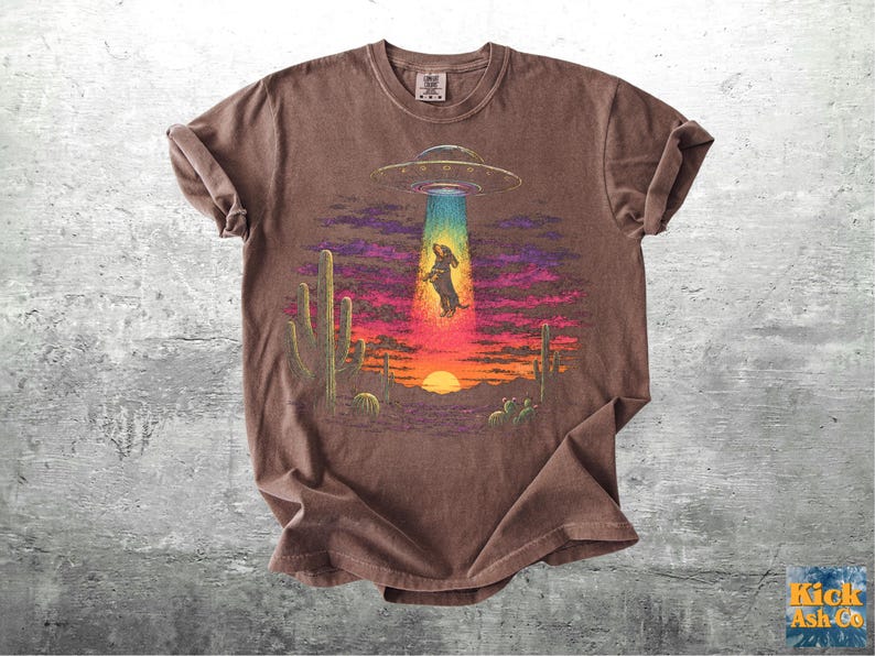 May include: A brown short-sleeve t-shirt featuring a graphic of a dachshund being abducted by a flying saucer in a desert landscape at sunset. The scene includes cacti, a bright sun on the horizon, and a colorful sky with purple and orange hues.